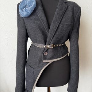 Può includere: Un blazer grigio scuro con un design asimmetrico unico, rifinito in grigio e con chiusura a un solo bottone. Una cintura borchiata stringe la vita e un fiore di tessuto blu adorna il risvolto. Il blazer è esposto su un manichino nero.