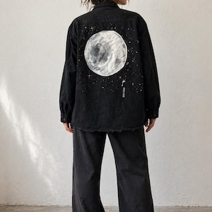 以下が含まれることがあります： 黒色のデニムジャケットとパンツのセット。ジャケットの背面には、手描きの大きな白い月のデザインと星のアクセントがあります。パンツはワイドレッグで、カジュアルでコーディネートされた服装です。