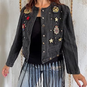 Può includere: Una giacca in denim grigio antracite con scollo rotondo, bottoni in metallo argentato e maniche lunghe. La giacca è decorata con varie toppe ricamate, tra cui stelle e stemmi, insieme a borchie in metallo argentato sparse.