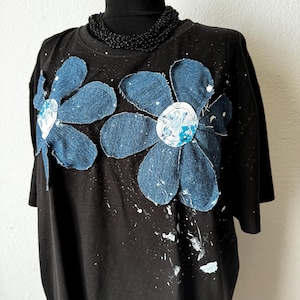 Può includere: T-shirt nero con due grandi applicazioni di fiori in denim con dettagli bianchi e blu e schizzi di vernice bianca. Una collana di perline nere è intorno al collo del manichino.