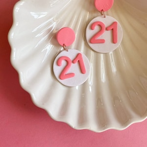 May include: A pair of pink and white dangle earrings with the number 21 in pink on a white circle.