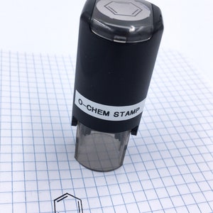 May include: A black O-CHEM STAMP with a clear base is shown on a white grid paper. The stamp has a hexagonal design on top and prints a benzene ring. The printed benzene ring is next to the stamp.