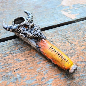 Puede incluir: Una pipa de cerámica con forma de cabeza de demonio con cuernos, pintada en negro, blanco, naranja y amarillo con un cuerpo a rayas amarillo y naranja y una serie de agujeros a lo largo del lado.