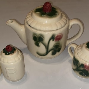 May include: A cream ceramic tea set featuring a teapot, sugar bowl, and salt and pepper shakers. Each item is adorned with a strawberry and shamrock motif. The teapot has a lid with a strawberry-shaped handle.