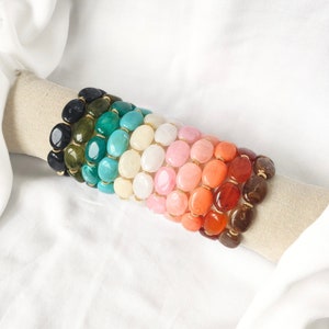 Puede incluir: Una pila de pulseras coloridas hechas con cuentas ovaladas. Las pulseras están dispuestas en un patrón de arcoíris, comenzando con negro y terminando con marrón.