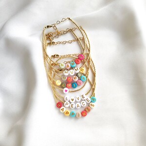 Peut inclure: Bracelets dorés avec des perles de lettres colorées épelant des mots tels que "MAMIE", "FAMILLE", "AMOUR", "PAIX" et "BONHEUR". Les bracelets ont des fermoirs en forme de cœur dorés, présentés sur une surface en satin blanc.