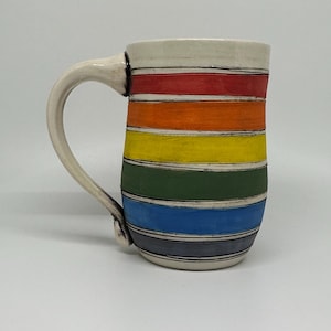 May include: A ceramic mug with a cream-colored handle and rim. The body of the mug is decorated with horizontal stripes in a rainbow of colors: red, orange, yellow, green, blue, and gray. The mug has a capacity of approximately 16 ounces.