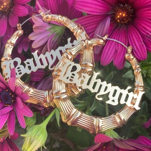 May include: Gold hoop earrings with a bamboo-style design. The earrings feature the word "Babygirl" in a bold, cursive font.