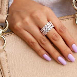 Może przedstawiać: Zbliżenie dłoni z pierścionkiem z różowego złota z wieloma rzędami przezroczystych, okrągłych kamieni. Pierścionek znajduje się na środkowym palcu dłoni. Dłoń spoczywa na jasnobrązowej skórzanej powierzchni.