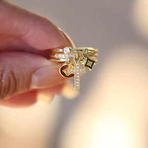 May include: A set of gold rings, designed to be stacked. The rings have a small diamond, a cross charm, a heart charm, and a star charm. The rings are made of gold and are intended to be worn together.