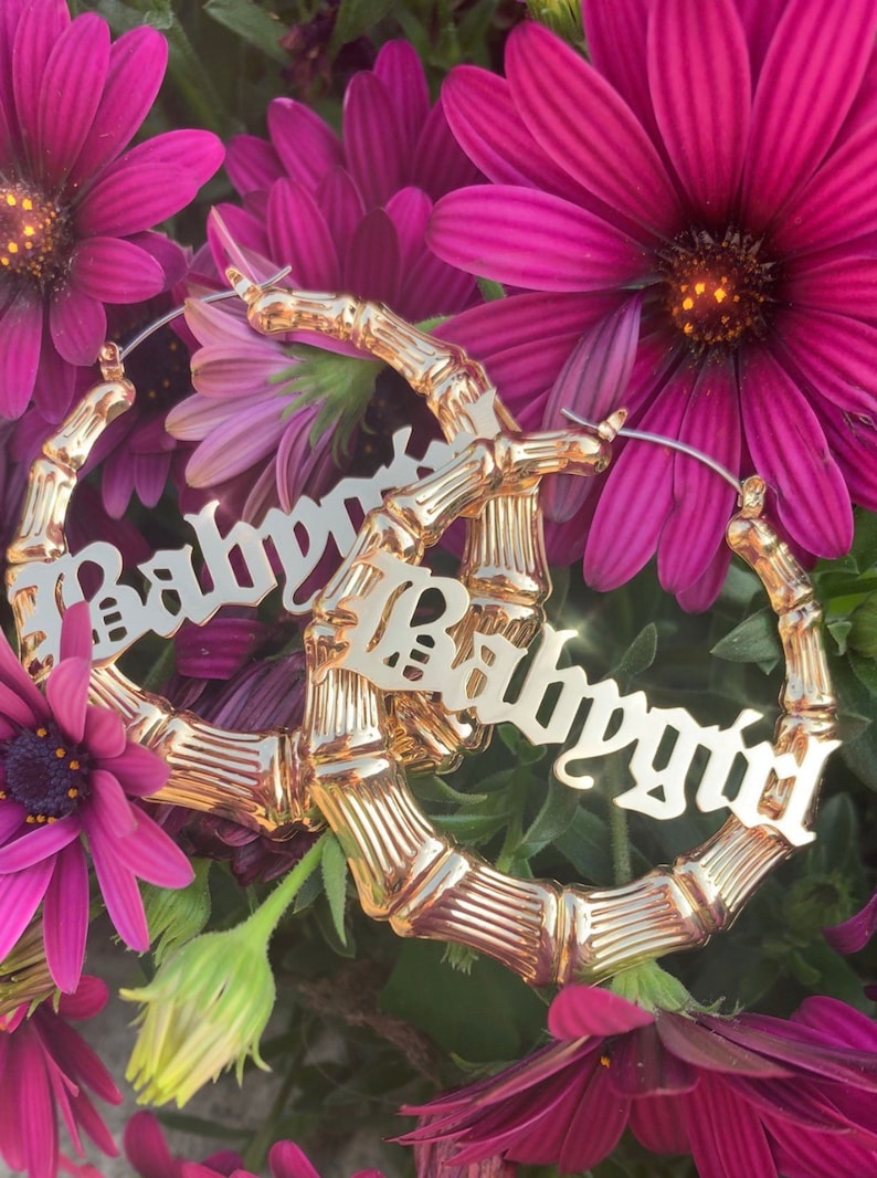 K&ouml;nnte beinhalten: Goldene Creolen mit Bambus-Design. Die Ohrringe zeigen das Wort "Babygirl" in einer fetten, kursiv geschriebenen Schrift.