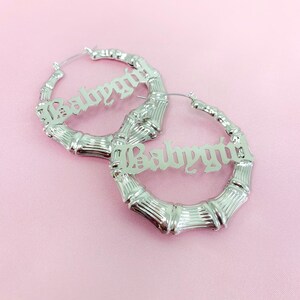 K&ouml;nnte beinhalten: Ein Paar silberne Creolen mit dem Wort "Babygirl" in gotischer Schrift. Die Ohrringe sind aus Bambusmaterial.
