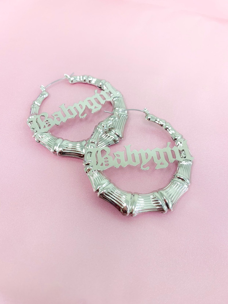 K&ouml;nnte beinhalten: Ein Paar silberne Creolen mit dem Wort "Babygirl" in gotischer Schrift. Die Ohrringe sind aus Bambusmaterial.