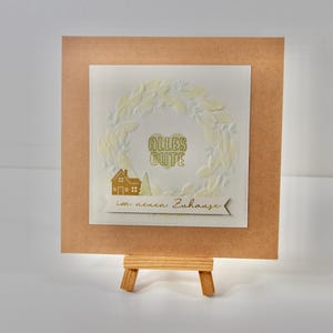 Könnte beinhalten: Eine Grußkarte mit braunem Rand und weißer Mitte. Die Mitte zeigt ein Kranzdesign, die Worte "Alles Gute" und eine Hausillustration. Die Karte wird auf einer kleinen Holzstaffelei präsentiert.