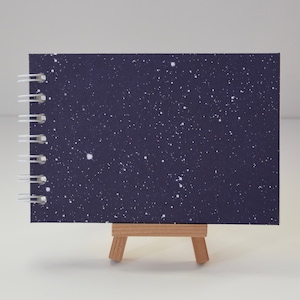 Può includere: Un taccuino a spirale con una copertina blu scuro punteggiata di punti bianchi, che ricorda un cielo stellato. Il taccuino è appoggiato su un piccolo cavalletto di legno, che ne mette in risalto il design. La rilegatura a spirale è bianca.