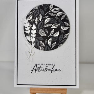 May include: A white card with a black border features a circular design of black and white leaves. The card includes the German text "AUFRICHTIGE Anteilnahme" and a decorative sprig. The card is displayed on a small wooden easel.