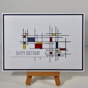 Pode incluir: Um cartão de felicitações branco com uma borda azul marinho e um design geométrico de linhas pretas e retângulos coloridos em vermelho, amarelo e azul. Uma faixa com o texto "HAPPY BIRTHDAY" está na parte inferior.