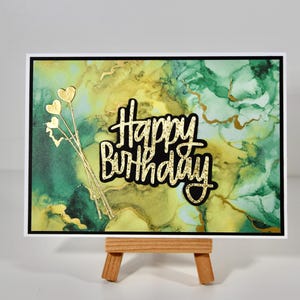 Peut inclure: Carte de vœux d'anniversaire avec un fond abstrait coloré vert et jaune. La carte présente des accents dorés, dont l'inscription "Happy Birthday" et des ornements en forme de cœur. Présentée sur un petit chevalet en bois.