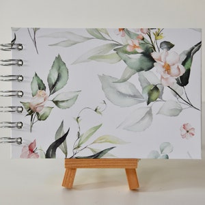 May include: A white spiral bound journal with a watercolor floral design featuring pink and green flowers and leaves. The journal is on a small wooden easel.