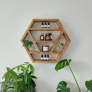 May include: A hexagonal wooden shelf with five shelves, featuring small glass bottles and jars on the shelves. The shelf is mounted on a white wall with green plants on either side.