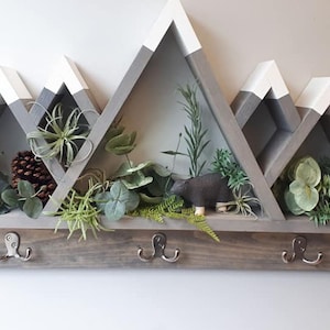 May include: A gray and white wooden mountain-shaped shelf with three hooks on the bottom. The shelf has three triangular compartments, each filled with artificial greenery. A small wooden bear figurine is in the middle compartment.