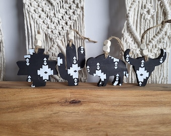 May include: Four black and white wooden ornaments with geometric patterns. The ornaments are shaped like a cactus, a buffalo, a skull, and a star. Each ornament has a white bead and twine for hanging.