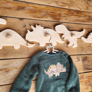Peut inclure: Ensemble de crochets muraux en bois en forme de dinosaures, accompagnés d'un sweat-shirt vert avec un appliqué de dinosaure. Les crochets sont en bois clair et sont fixés sur un fond de planches de bois.