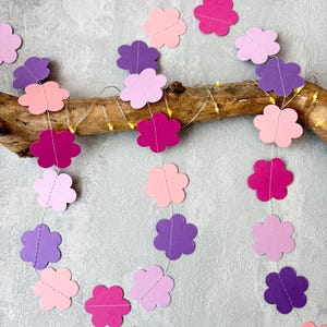Könnte beinhalten: Eine dekorative Girlande mit mehreren Papierblumen-Ausschnitten in Rosa-, Magenta- und Violetttönen, die auf einen dünnen weißen Faden aufgezogen sind. Die Girlande ist über ein Stück natürliches Treibholz drapiert, das von kleinen Lichterketten beleuchtet wird.