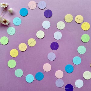 Könnte beinhalten: Eine Girlande aus bunten Papierkreisen in Pastelltönen von Gelb, Grün, Blau, Rosa und Lila, auf einem dünnen weißen Faden aufgereiht. Die Girlande ist in einem wellenförmigen Muster auf einem lila Hintergrund angeordnet, mit Trockenblumen.