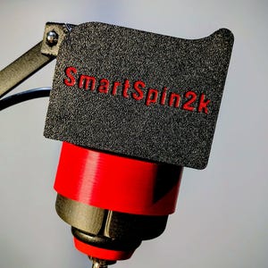 May include: A black textured rectangular component with the red text SmartSpin2k printed on the front. This piece is attached to a red and black cylindrical base with a metallic rod extending from the bottom, featuring a small metal hinge.