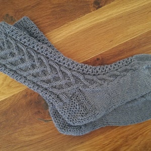 Könnte beinhalten: Ein Paar graue Strickstrümpfe mit Zopfmuster am oberen Teil. Die Socken sind aus einem weichen Material gefertigt und liegen auf einer Holzoberfläche.
