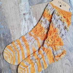 Könnte beinhalten: Ein Paar handgestrickte Socken in Orange- und Grautönen mit Streifen- und Lochmuster. Die Socken werden auf einer Holzoberfläche präsentiert. Das Etikett oben lautet "TAIOA".