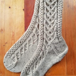Könnte beinhalten: Ein Paar hellgraue Strickstrümpfe mit Zopfmuster. Die Socken sind aus weichem, gesprenkeltem Garn gefertigt und haben ein strukturiertes Muster. Die Socken werden auf einer Holzoberfläche präsentiert.