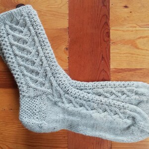 Könnte beinhalten: Ein Paar hellgraue Strickstrümpfe mit Zopfmuster. Die Socken sind aus einem weichen, strukturierten Garn gefertigt und weisen ein detailliertes Muster auf der Vorderseite und Oberseite auf. Die Socken liegen auf einer Holzoberfläche.