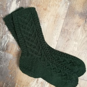 Könnte beinhalten: Ein Paar dunkelgrüne Strickstrümpfe mit einem strukturierten Zopfmuster. Die Socken sind aus einem weichen, warmen Material gefertigt. Die Socken liegen auf einer Holzoberfläche.