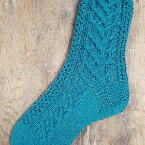 Könnte beinhalten: Eine einzelne, türkis gestrickte Socke. Die Socke hat ein Zopfmuster auf der Vorderseite und ein strukturiertes Muster an den Seiten. Ferse und Zehen sind einfarbig türkis. Die Socke wird auf einer Holzoberfläche präsentiert.