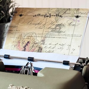 May include: An olive green Smith-Corona typewriter with a sheet of paper inserted, displaying vintage script and floral illustrations. The paper is held in place by the typewriter's rollers. A potted plant sits in the background.