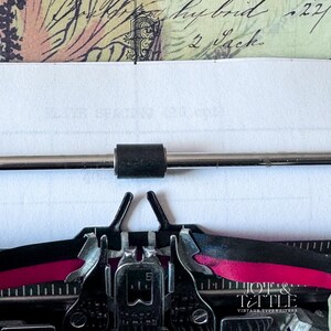 May include: Close-up of a vintage typewriter with a sheet of paper inserted. The typewriter's keys and ribbon are visible, with the ribbon showing a vibrant pink color. The paper has handwritten text and is partially visible.
