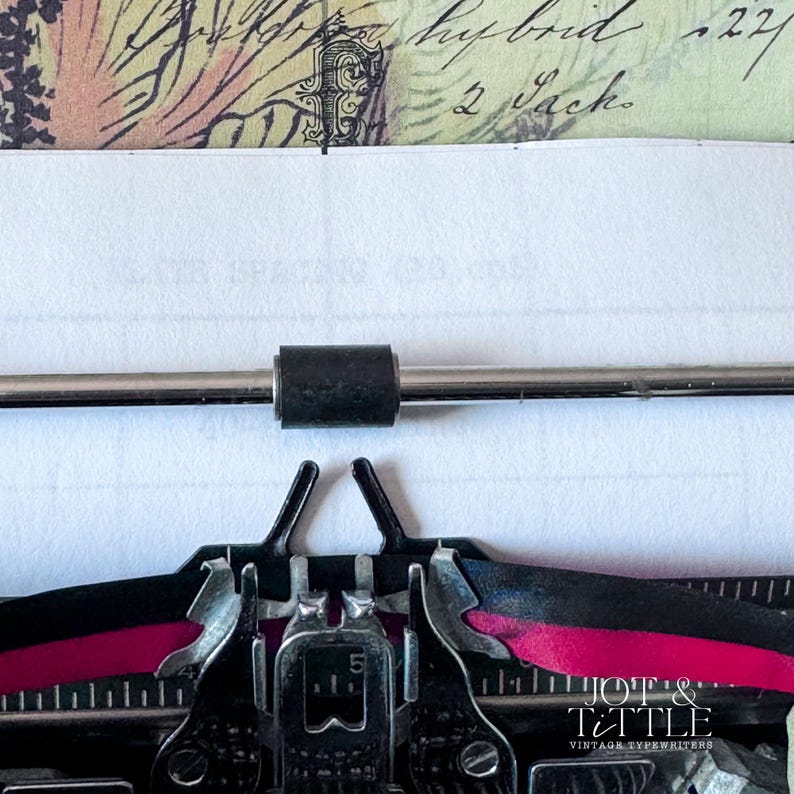 May include: Close-up of a vintage typewriter with a sheet of paper inserted. The typewriter's keys and ribbon are visible, with the ribbon showing a vibrant pink color. The paper has handwritten text and is partially visible.
