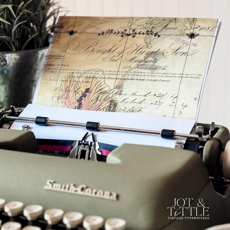 May include: An olive green Smith-Corona typewriter with a sheet of paper inserted, displaying vintage script and floral illustrations. The paper is held in place by the typewriter's rollers. A potted plant sits in the background.