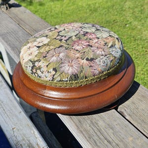 May include: A round, wooden pincushion with a floral tapestry top. The cushion features a variety of flowers in shades of pink, green, and cream against a black background. The wooden base is a rich, dark brown.
