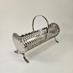 May include: A silver-toned metal basket with a long, rectangular body featuring an intricate lattice pattern. The basket has a scalloped shell design at both ends, a single arched handle, and stands on four small, curved metal feet.