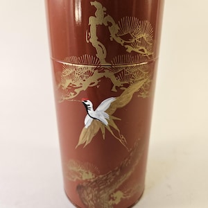 May include: A cylindrical reddish-brown tea canister featuring a gold-painted pine tree design and a white crane with a red crest in flight. The container has a smooth, glossy finish and a horizontal seam near the top indicating a removable lid.