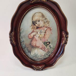 May include: An oval-shaped picture frame with a dark burgundy finish and gold-toned decorative accents. The frame contains a print of a young child with blonde hair wearing a pink patterned top, holding a small doll in a grassy outdoor setting.