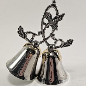 May include: A pair of silver-colored bells with gold-colored rims, connected by a decorative, leafy metalwork. The bells are polished and reflect the surrounding light. The design includes a small ball at the top.