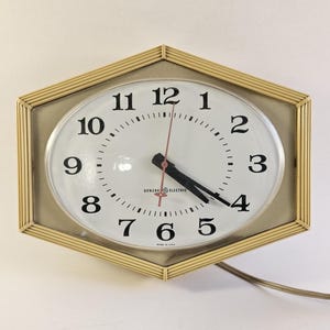 May include: Vintage wall clock with an oval face and a geometric frame. The clock face is white with black numerals and hands, and a red second hand. The frame is a light beige color. The clock reads "General Electric" at the bottom.