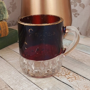 May include: A vintage glass mug with a ruby red and clear diamond-cut design. The mug has a gold rim and handle. A bouquet of pink and orange gerbera daisies sits in a copper vase in the background.