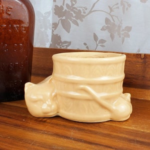 May include: A ceramic planter shaped like a cat with a barrel on its back. The planter is a light peach color and has a textured surface. The planter is sitting on a wooden surface.