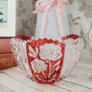 May include: A white picture frame with a quote about hope and a floral design with birds.  The frame is sitting on top of a stack of three pink books.  A clear glass bowl with a red rim and a floral design is on the right side of the image.  A white vase with a pink ribbon is filled with pink and white roses.