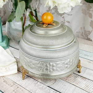 May include: A vintage silver music box with a floral design and gold accents. The music box has a round lid with a knob on top and sits on four gold feet. The music box is on a white wooden surface with a bouquet of white and peach flowers in a glass vase.